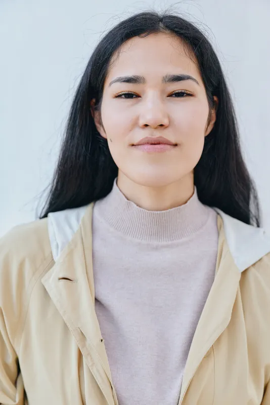 Close up of female with calm expression wearing beige coat and light sweater against neutral background, soft lighting and casual urban fashion vibe, stylish look
