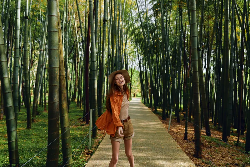 Woman walking on a bamboo path in a forest wearing a wide hat and smile, casual shorts and jacket, summer nature scene with sunlight filtering through tall green stalks.