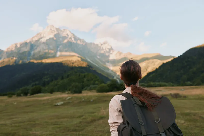 A young woman with a backpack faces distant snowy peaks in a wide valley, embodying a moment of adventure and exploration.