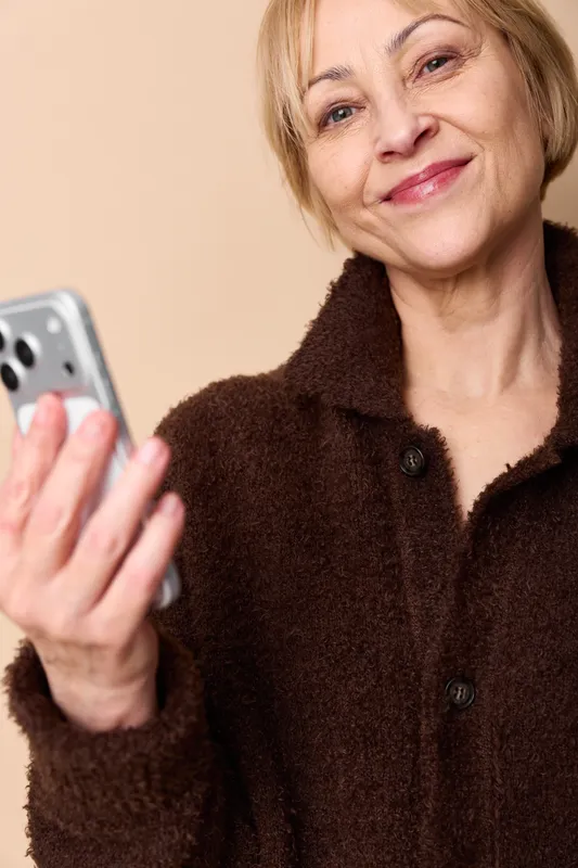 Woman with smartphone portrait, smiling selfie, senior female in brown sweater, holding phone with hand, indoor beige background, technology communication