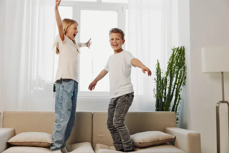 Two happy children jumping on a beige sofa in bright living room with natural light and modern decor. Concept of childhood fun, energy, and playful indoor activity.
