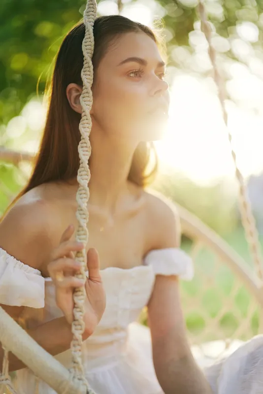 A serene young woman in a white dress sits on a swing, capturing sunlight in an outdoor setting, evoking a sense of tranquility and summer bliss