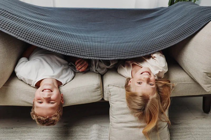 Two children lie upside down on a beige sofa covered by a gray blanket, smiling and playing indoors on a cozy carpet. Concept of childhood fun, family bonding, and home comfort.