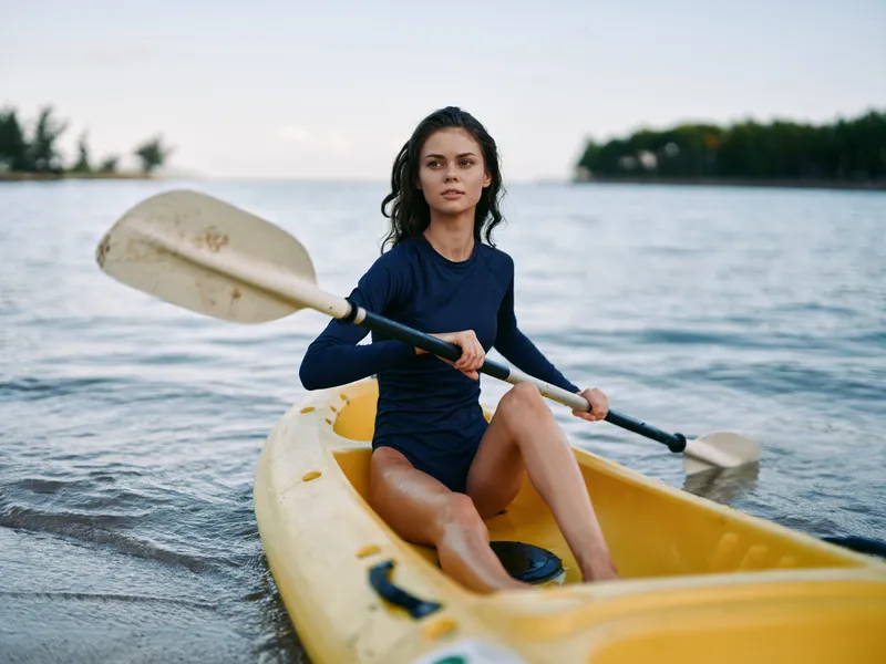 Young woman sits in a yellow kayak, holding a paddle across calm turquoise water, wearing a dark long sleeve swimsuit, ready for a peaceful coastal outing at golden hour.