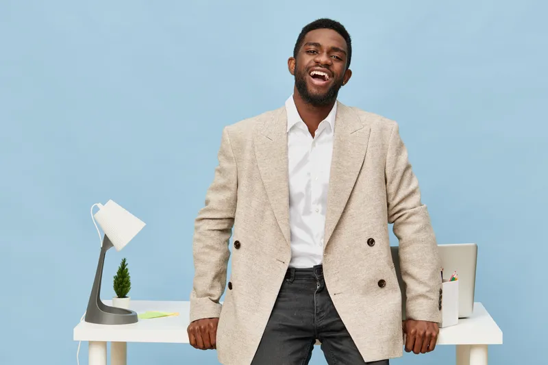 Professional confident businessman standing in modern office with white desk, smiling broadly, dressed in beige blazer and white shirt, showcasing success and positivity, against plain blue background