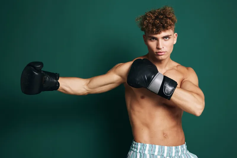 Young man with curly hair wearing boxing gloves and striped shorts practicing punch against green background. Athletic shirtless male training for fitness and combat sport indoors.