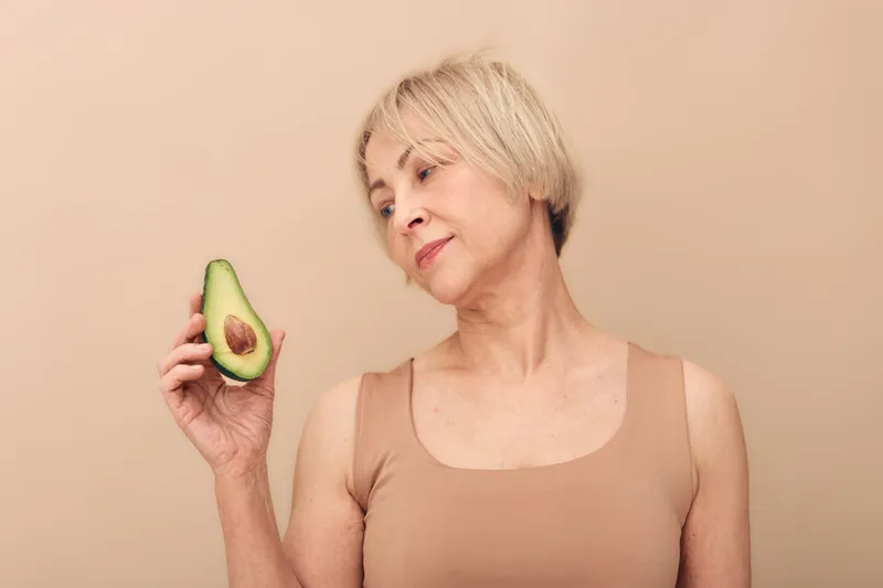 Mature woman with short blond hair holds half of avocado, looking at it thoughtfully on beige background. Concept of healthy lifestyle, nutrition, and natural diet for aging skin.