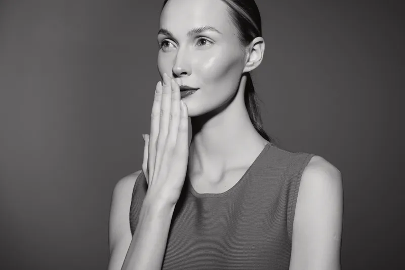 White woman with fair skin covers mouth with hand, looking sideways against plain background. Concept of secrecy, emotion, and expression.