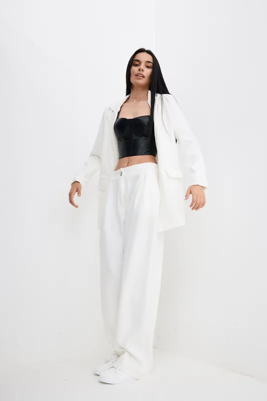 Young woman in white oversized pants and blazer with black leather corset top posing confidently against minimalistic white background, showcasing modern fashion style.