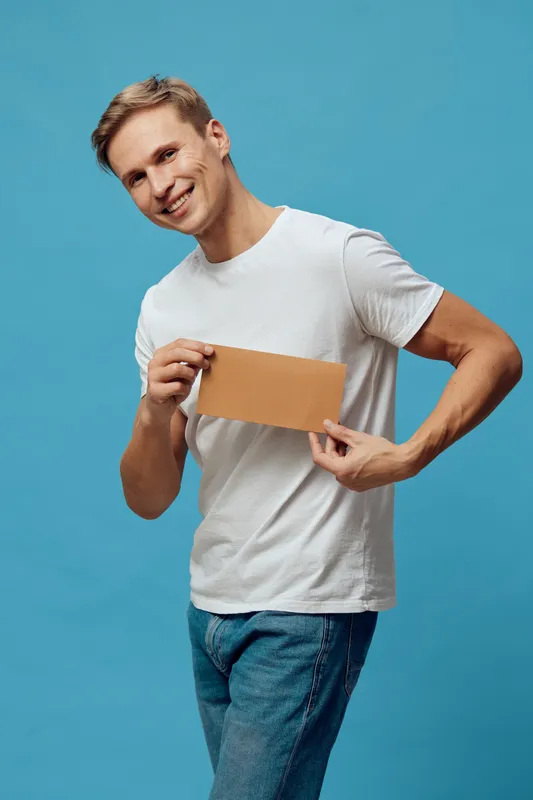 Smiling young man with blond hair holding a blank cardboard sign while posing on a bright blue background. Casual attire, cheerful mood, lifestyle, studio shot, vibrant colors, positive expression, yo
