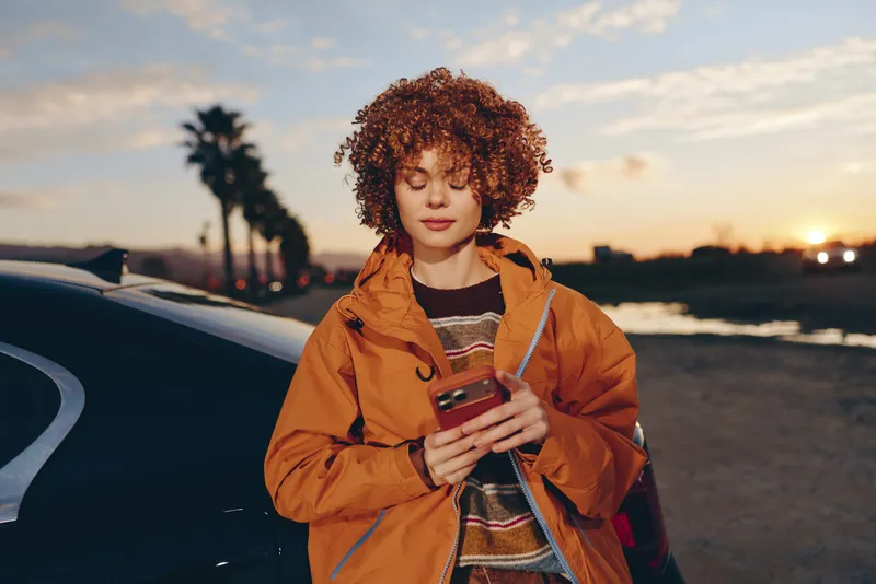 Woman lifestyle outdoors at sunset holding a smartphone beside a parked car on a coastal road, wearing a brown jacket, curly hair, soft breeze, warm sky, relaxed smile