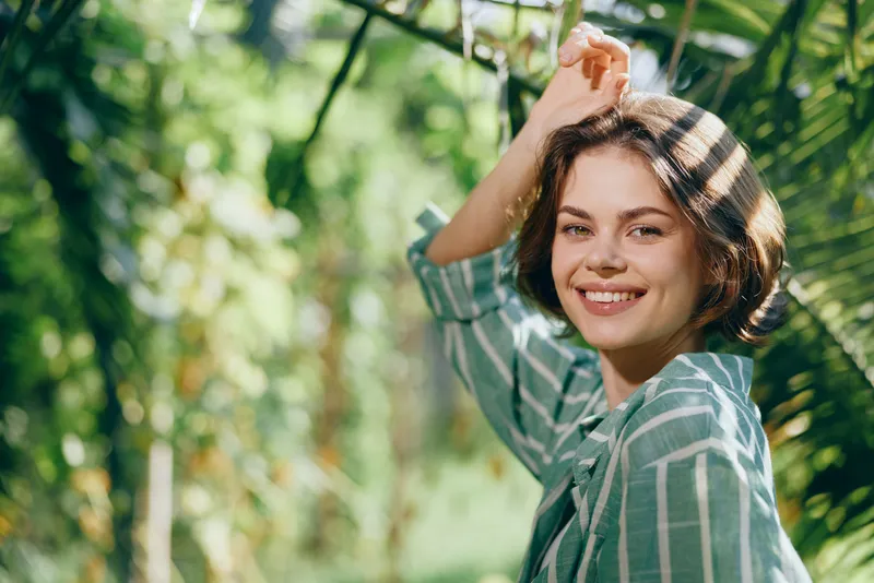 Portrait of a young woman outdoors, smiling softly while lifting one arm under dappled sunlight, wearing a striped green shirt.