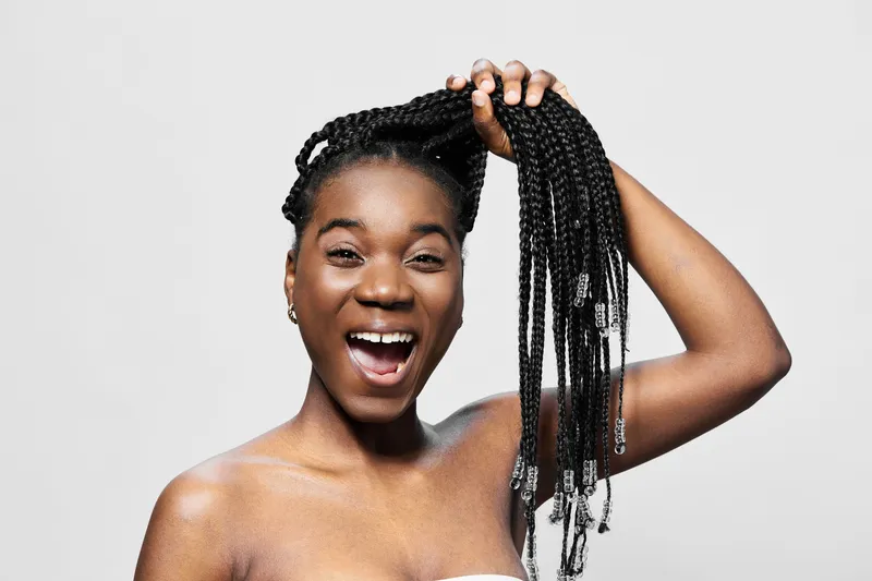 Joyful African woman with braids showcasing her hair in a bright studio, expressing confidence and happiness with a playful pose and neutral background