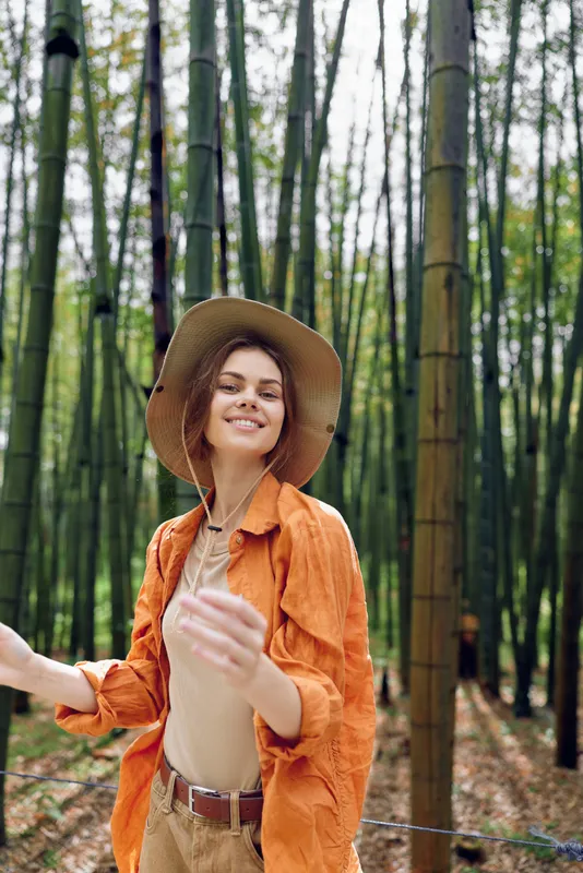 Woman in a bamboo forest wears a hat and smile, nature portrait outdoors with green trees and casual fashion, young female enjoying travel, sunlight and serene woodland leisure