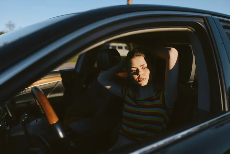 Woman inside a car, relaxed and thoughtful, sunlight across her face, striped shirt, window open, warm retro vibe, candid lifestyle moment, natural colors