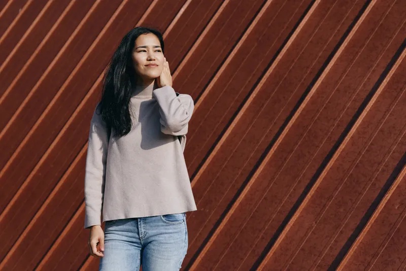 Woman in casual sweater and jeans posed thoughtfully against a diagonal wooden wall, outdoor setting, natural light, simple composition conveying calm urban mood