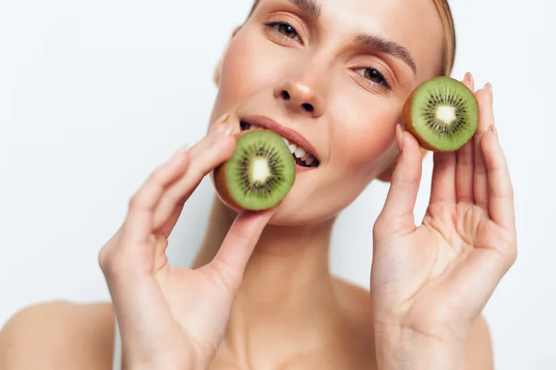 A smiling woman holds two halves of a fresh kiwi fruit near her face against a white background. The image conveys health, natural beauty, and fresh organic lifestyle concepts.