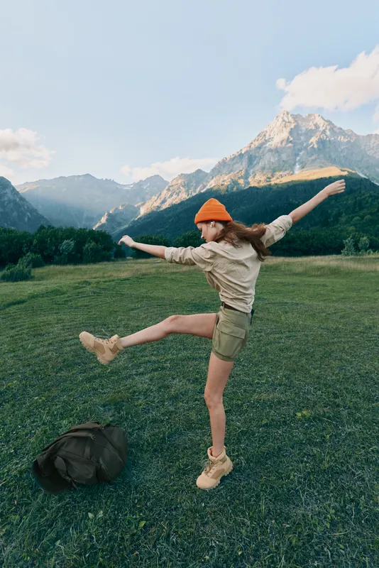 Young woman wearing an orange beanie jumps with arms outstretched across a grassy mountain meadow, backpack resting nearby, dramatic alpine peaks in the background.