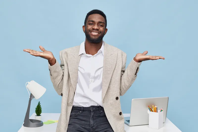 Smiling young African American man with beard, wearing casual beige blazer and white shirt, standing in office environment with desk, laptop, and stationery, demonstrating a relaxed and confident mood