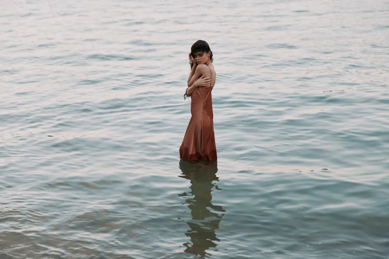 A lone figure stands waist deep in calm water, wearing a long rust dress. Soft light highlights gentle ripples, creating a tranquil, introspective mood.