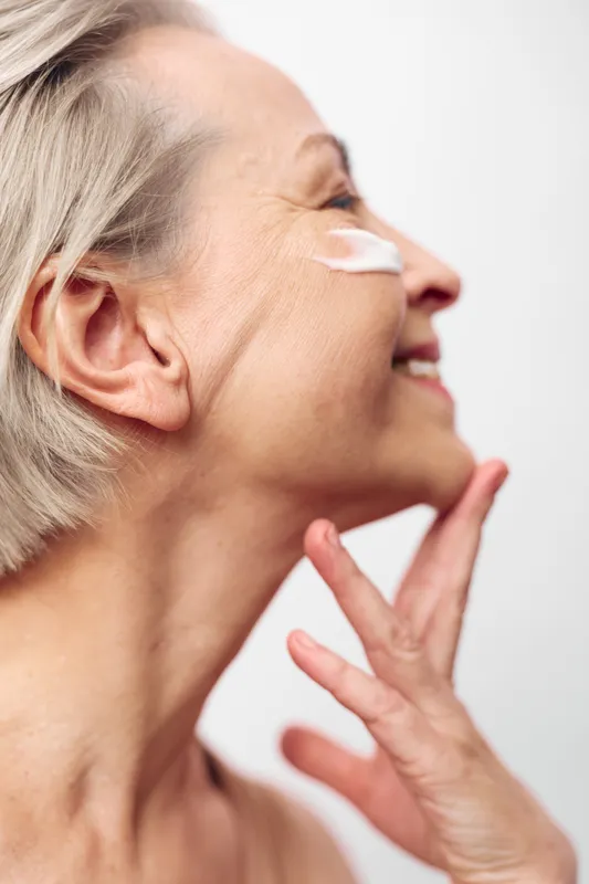 Senior woman with short hair applies moisturizing cream on her cheek, smiling gently with closed eyes. Concept of skincare, aging beauty, self-care, and healthy skin routine.