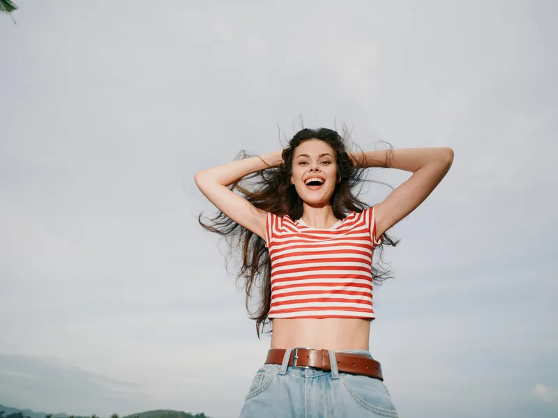 Bright, joyful portrait of a young woman standing outdoors, wearing a striped crop top and jeans, smiling with hands in her hair, capturing carefree summer energy and confident spontaneity against a