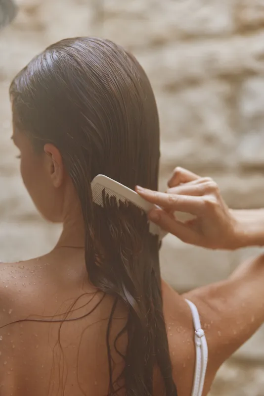 Woman combing wet hair with white comb outdoors, smooth brown hair, natural light, skin care routine, calm atmosphere, summer setting