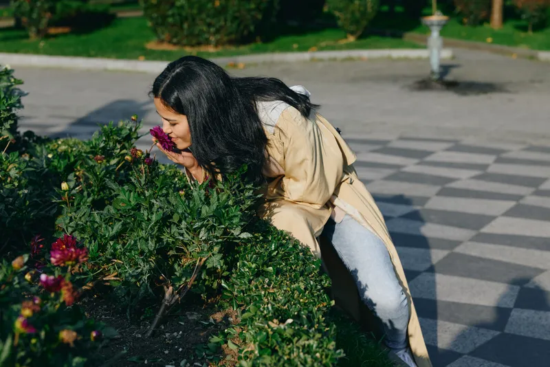 Flowers, fragrance, outdoor garden scene with a figure leaning toward lush greenery, savoring scent in a park setting, natural light, casual coat, calm mood, soft breeze, quiet moment.