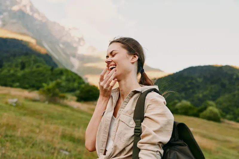 A happy young woman with a backpack laughs outdoors in a mountainous landscape during daytime. She wears casual clothes and enjoys nature, representing lifestyle and adventure.