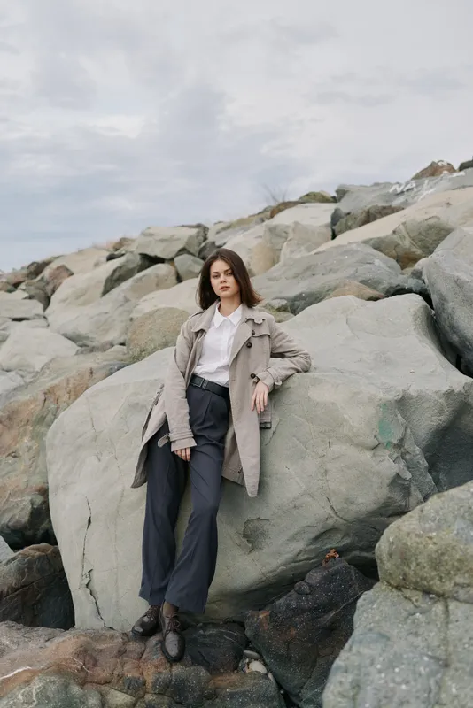 Young woman in casual outfit poses leaning on large rocks outdoors under cloudy sky. Natural scenery with rocky background creates calm and contemplative mood for fashion or lifestyle concept.