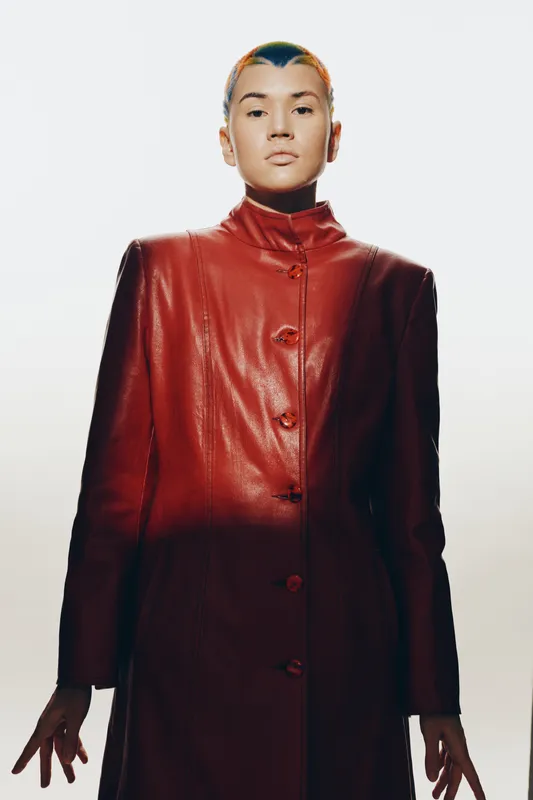 Stylish woman in a red leather coat with a modern haircut, standing confidently against a plain white background, showcasing bold fashion and minimalistic elegance.