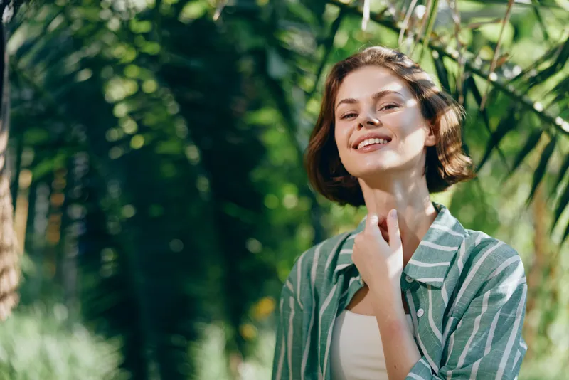 A cheerful woman stands outdoors in bright daylight, smiling with relaxed confidence. She wears a striped shirt, enjoying nature and sunshine. The scene conveys freshness, warmth, and optimism,
