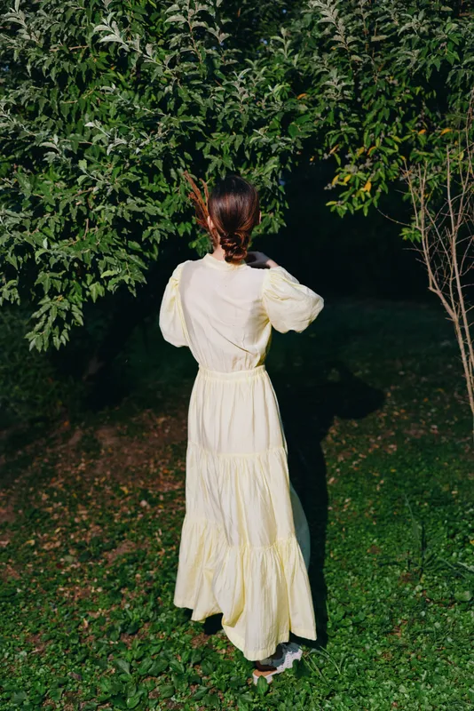 Woman in a pale yellow dress stands in a lush garden at night, seen from behind, strolling along a dimly lit path among trees and greenery, with soft shadows and a tranquil mood.