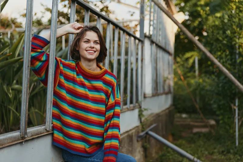 Woman in a rainbow sweater smiling and leaning against a greenhouse glass, casual outfit with short curly hair, 20s model enjoying relaxed outdoor lifestyle portrait in a garden.