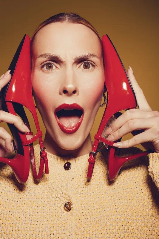 Surprised woman with red lipstick holds red high heel shoes near face, wearing yellow textured jacket. Concept of fashion, style, beauty, and bold makeup in studio lighting.