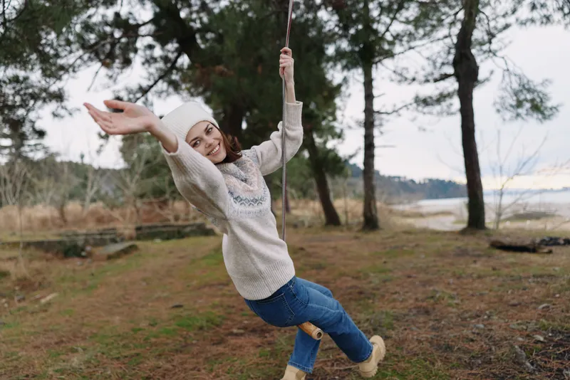 Woman on a wooden swing in a park, wearing a knit sweater and jeans, smiling as she swings, trees in the background, autumn light, carefree moment in nature, outdoor joy
