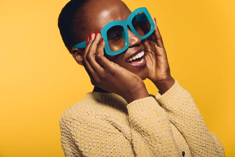 Happy dark skinned African woman wearing oversized blue sunglasses and beige sweater smiles with joy on yellow background. Concept of fashion, summer style, and positive emotions.