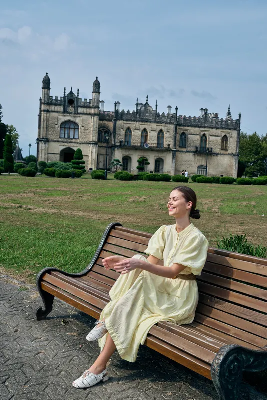 Woman in a light yellow dress sits on a wooden bench in an outdoor park with a historic castle in the background, enjoying a peaceful moment in daylight.