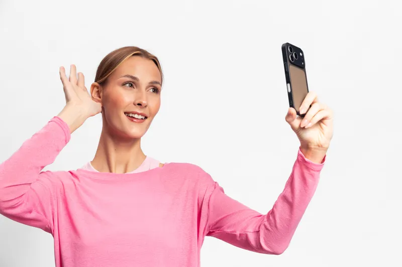 Young woman in pink sweater takes selfie with smartphone on white background. She smiles and poses with hand near ear. Concept for social media, communication, and modern technology.