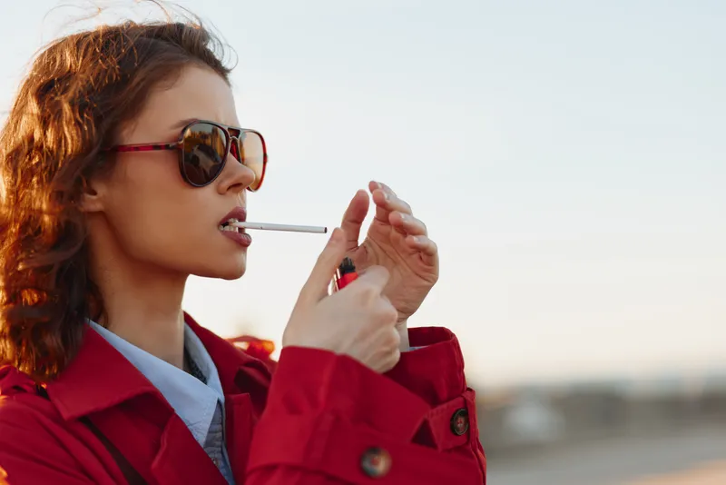 Young woman with curly hair wearing sunglasses and a red coat lights a cigarette outdoors during a clear day with soft natural light.