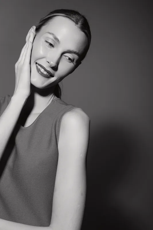 Young Caucasian woman with light skin smiles gently, touching face with hand. Studio portrait with soft lighting and dark background. Concept of beauty and skin care.