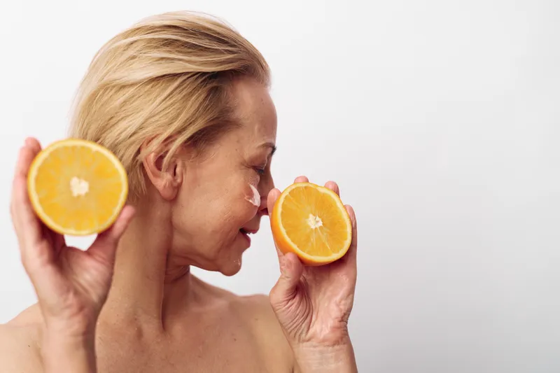 Blond woman with bare shoulders holds two halves of fresh orange near face, eyes closed, applying natural skincare cream. Concept of organic beauty, wellness, and healthy lifestyle with citrus fruit.