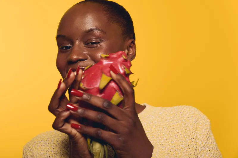 Smiling dark skinned African woman holds a ripe dragon fruit close to her face against a yellow background. Concept of healthy lifestyle, fresh fruit, natural beauty, and wellness.