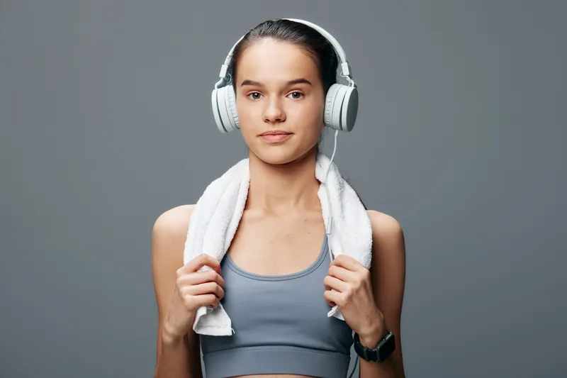 Woman in fitness gear with headphones and a towel around her neck, ready for a workout. Confident expression, simple studio backdrop, athletic routine and music guiding her active lifestyle.