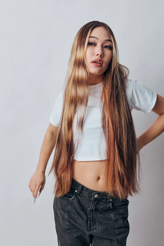 Young woman with long straight hair wearing a white crop top and black jeans posing confidently with hand on hip in front of light gray background.