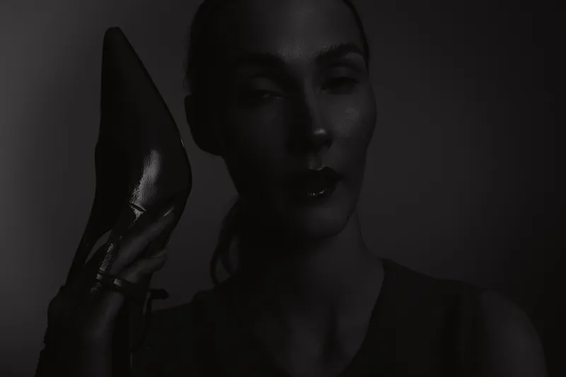 Portrait of a dark skinned African woman holding a high heel shoe near her face in a low light setting. Concept for fashion, elegance, beauty, and style.