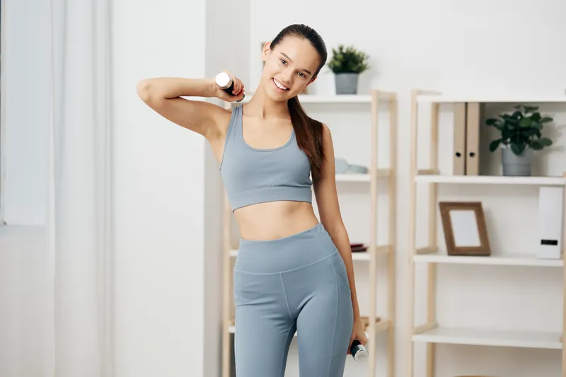 A woman in athletic wear performs a shoulder press with light dumbbells at home, bright room with shelves and plants, conveying strength, focus and a positive fitness routine during a personal