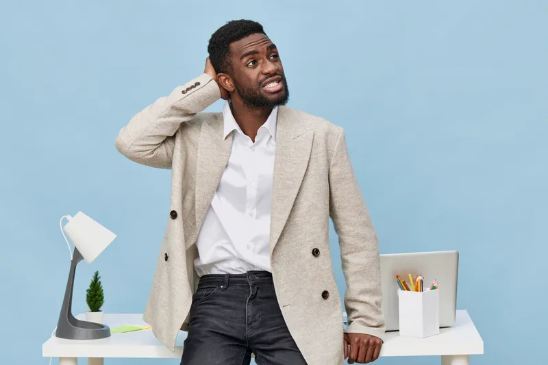 Disappointed confused young African American man in casual beige blazer and white shirt looks unsure at workspace with laptop and stationery on light blue background, expressing frustration or confusi