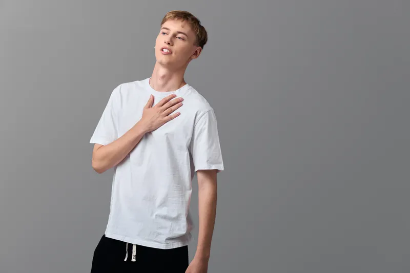 Man in white t shirt places hand on chest, standing against a neutral backdrop, conveying surprise, emotion, and vulnerability in a simple studio pose.