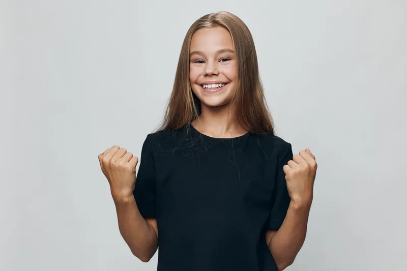 Smiling girl with long hair wearing black shirt showing fists in celebration or success gesture on light background. Child with happy expression and confident posture in studio.